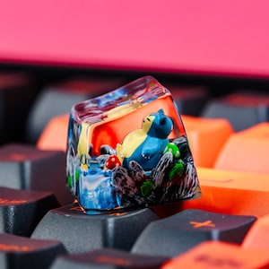 Puede incluir: Un keycap de resina transparente con un personaje de dibujos animados azul y amarillo en su interior. El personaje está sentado en una cama de resina verde y marrón. El keycap está sobre un teclado negro.