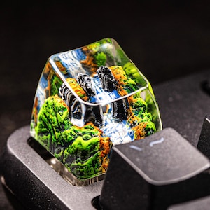 Könnte beinhalten: Eine individuelle Tastaturkappe mit einer Miniatur-Landschaftsszene. Die transparente Kappe zeigt ein detailliertes Diorama aus grünen Bäumen, einem Wasserfall und schwarzen Felsformationen. Die Kappe ist auf einer schwarzen Tastatur montiert.