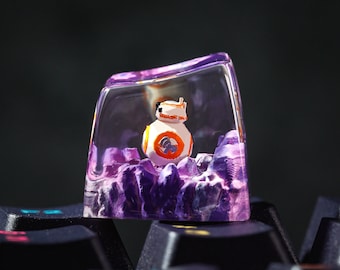 May include: A clear purple resin keycap with a small white and orange robot figurine inside. The keycap is sitting on a black keyboard.