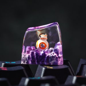May include: A clear purple resin keycap with a small white and orange robot figurine inside. The keycap is sitting on a black keyboard.
