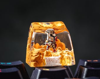 May include: A custom keycap featuring a miniature figure of a character from Star Wars, encased in a clear, amber-colored resin. The keycap is mounted on a black keyboard, with the Star Wars logo visible. The scene depicts a desert landscape.