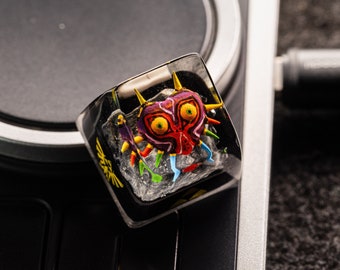 May include: A clear resin keycap with a colorful, detailed illustration of a video game character's mask. The mask is red, yellow, green, and purple with black eyes and a sharp, pointed design.