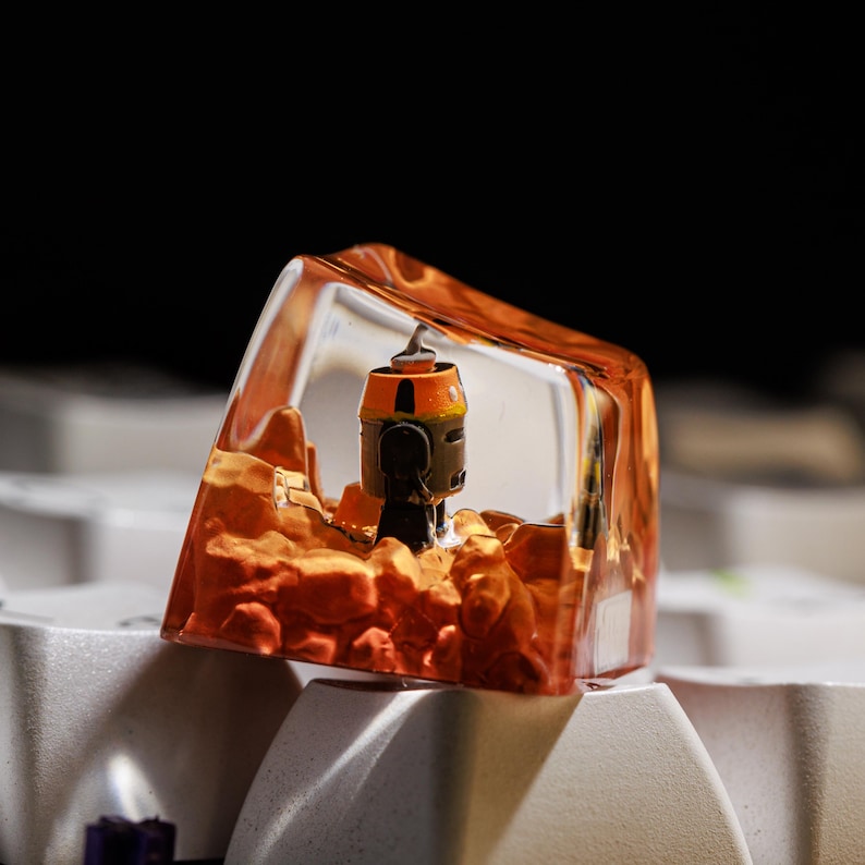 Puede incluir: Un keycap de resina transparente con una estaci&oacute;n espacial en miniatura de color naranja y negro en su interior. El keycap est&aacute; colocado sobre un teclado blanco.