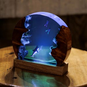 May include: A round, wooden base with a clear resin insert depicting a shark swimming in a blue ocean scene. The resin is illuminated from behind, creating a glowing effect. Two small figures of divers are also visible in the resin.