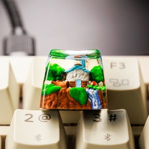 May include: A custom keycap featuring a miniature landscape with a house, trees, and a waterfall. The keycap is clear with a green rim and sits atop a computer keyboard. The keycap is a decorative item for computer enthusiasts.
