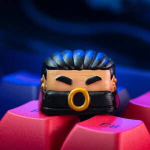 Pode incluir: Um keycap preto e dourado com um rosto de desenho animado usando uma m&aacute;scara preta. O keycap tem um anel dourado em volta da boca e cabelo preto.