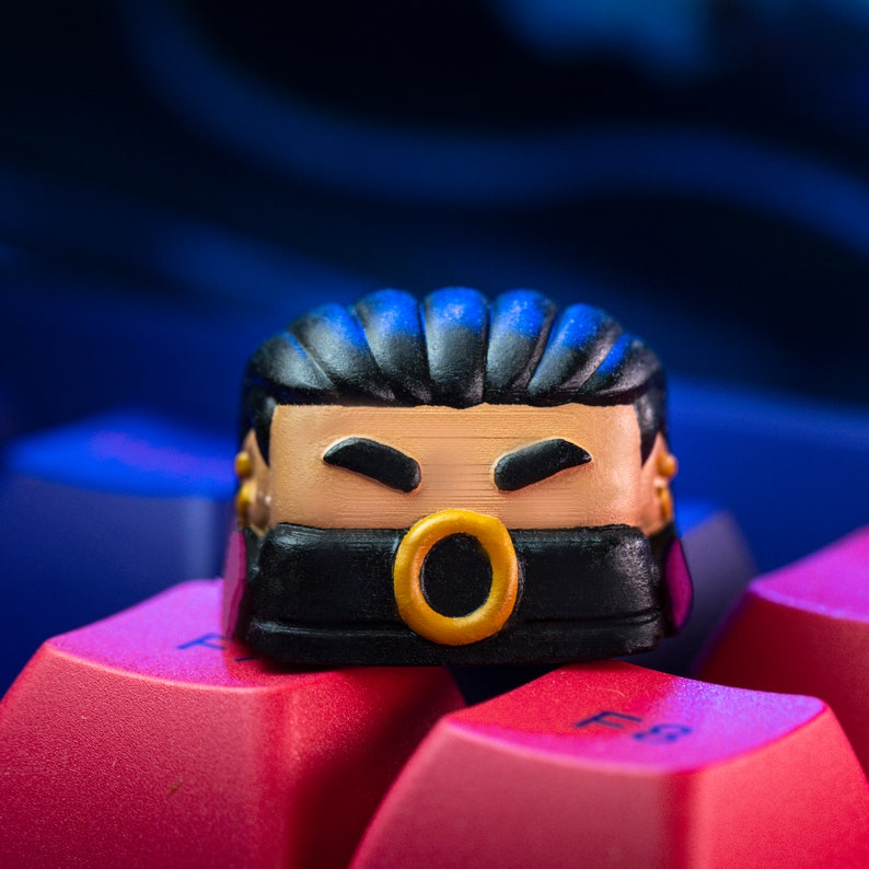 Pode incluir: Um keycap preto e dourado com um rosto de desenho animado usando uma m&aacute;scara preta. O keycap tem um anel dourado em volta da boca e cabelo preto.