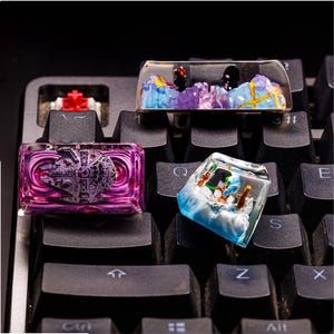 May include: Three custom keyboard keycaps with different designs. The first keycap is purple and features a spaceship. The second keycap is blue and features a winter scene. The third keycap is purple and features a mountain scene.