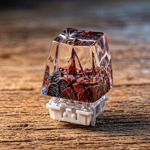 May include: A clear resin mechanical keyboard keycap featuring a miniature figure with a triangular head standing on a textured, dark red and black rocky landscape. The keycap is mounted on a white plastic mechanical switch base.