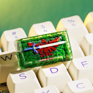 Può includere: Un copritasto in resina verde trasparente con un design a spada e scudo rosso e blu. Il copritasto è su una tastiera bianca con le lettere W, S, D, F e C visibili.