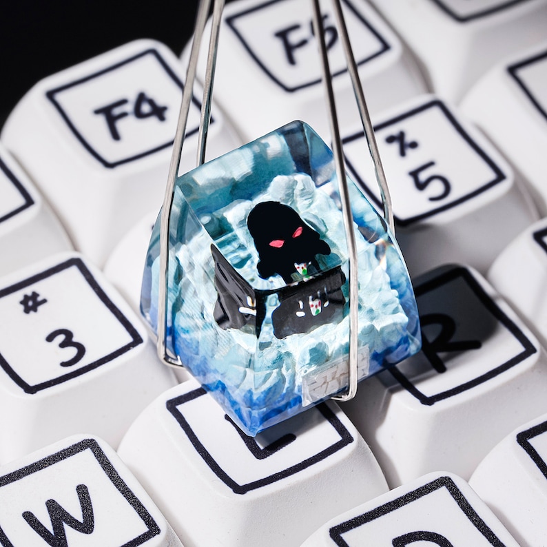 Puede incluir: Un keycap de resina azul y blanco con un dise&ntilde;o de monstruo negro y rojo. El keycap est&aacute; suspendido en un soporte de pl&aacute;stico transparente.
