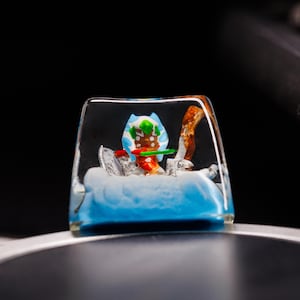 Puede incluir: Un keycap de resina transparente con una peque&ntilde;a figura de hombre de jengibre en su interior. La figura lleva un sombrero azul y blanco, una bufanda verde y un bast&oacute;n de caramelo rojo y blanco. La resina es azul y blanca, parecida a la nieve.
