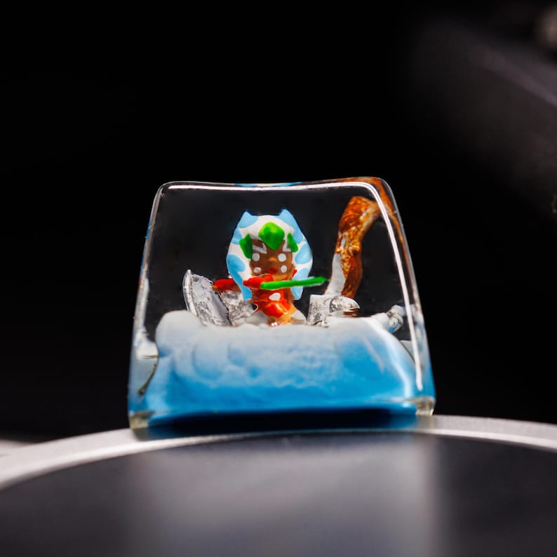 Puede incluir: Un keycap de resina transparente con una peque&ntilde;a figura de hombre de jengibre en su interior. La figura lleva un sombrero azul y blanco, una bufanda verde y un bast&oacute;n de caramelo rojo y blanco. La resina es azul y blanca, parecida a la nieve.