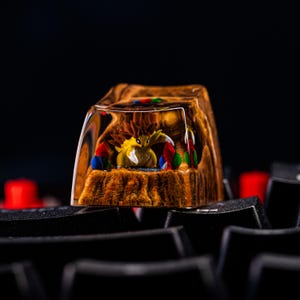 May include: A custom artisan keycap with a yellow creature and brown details, encased in clear resin. The keycap has a brown, textured base with red, blue, and green accents. The keycap is set on a black keyboard.