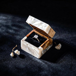 May include: A square ring box with a white and brown marbled resin exterior and a black velvet interior lining. A silver-toned ring with a single clear round-cut stone is centered inside the open box, accompanied by two small white dried flowers.