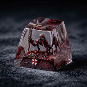Puede incluir: Una tecla artesanal de resina transparente que contiene una criatura miniatura roja y marrón con el cerebro expuesto y una lengua larga. La base incluye un pequeño logotipo en forma de paraguas de colores rojo, blanco y negro.