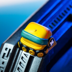 Pode incluir: Um rob&ocirc; de brinquedo amarelo e verde com uma cruz branca na cabe&ccedil;a. O rob&ocirc; est&aacute; sentado em uma superf&iacute;cie preta e prateada.
