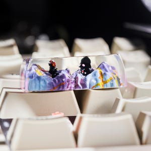 Puede incluir: Un keycap de resina transparente con un dise&ntilde;o de paisaje morado, azul y amarillo. Dos figuras negras son visibles dentro de la resina. El keycap est&aacute; sentado en un teclado blanco.