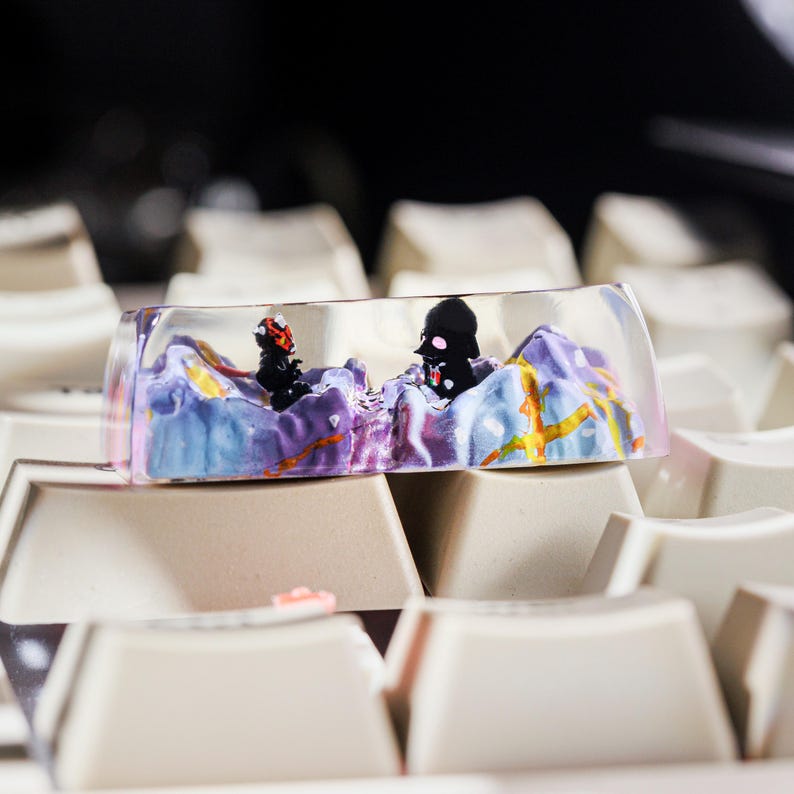 Puede incluir: Un keycap de resina transparente con un dise&ntilde;o de paisaje morado, azul y amarillo. Dos figuras negras son visibles dentro de la resina. El keycap est&aacute; sentado en un teclado blanco.