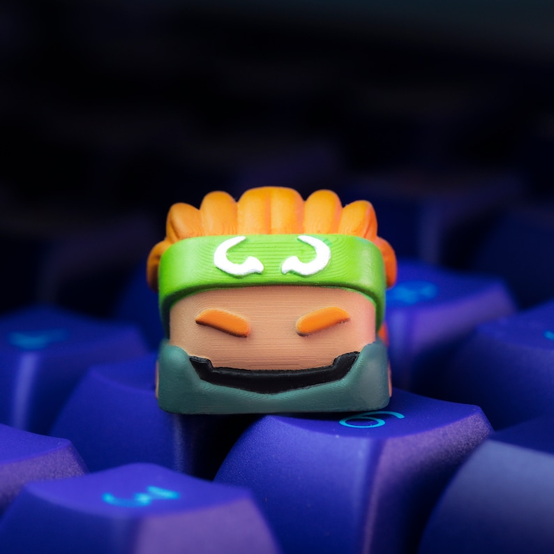 Pode incluir: Um keycap impresso em 3D verde, laranja e marrom com um rosto de desenho animado e uma faixa de cabe&ccedil;a branca. O keycap est&aacute; sentado em um teclado azul.