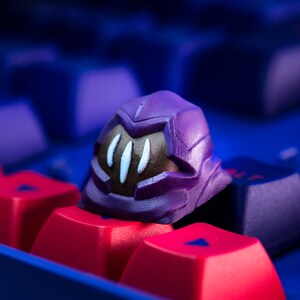 Pode incluir: Um keycap roxo e marrom impresso em 3D em forma de capacete com detalhes brancos. O keycap est&aacute; em cima de um teclado vermelho e azul.
