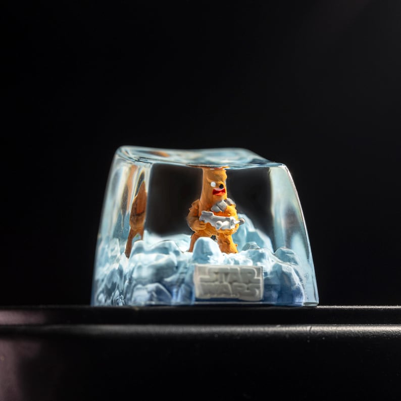 Puede incluir: Una peque&ntilde;a figurita naranja de un personaje de Star Wars est&aacute; encerrada en un cubo transparente parecido al hielo. El cubo est&aacute; sobre una superficie negra. La figurita sostiene un arma.