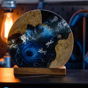 May include: A wooden base with a round resin top featuring a swirling blue nebula with two astronauts floating in space. The resin is set against a natural wood background.