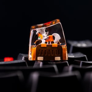 May include: A custom artisan keycap with a fox and a wolf encased in clear resin. The keycap has a brown and orange base, set on a black keyboard. The fox has orange and white fur, and the wolf is white and grey.