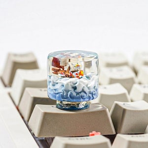 Puede incluir: Una tecla artesanal cilíndrica de resina que presenta un barco pirata en miniatura con una bandera de calavera navegando sobre olas esculpidas en azul y blanco. La tecla está montada sobre un interruptor de teclado mecánico de color beige.