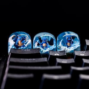 May include: Three custom keycaps with blue and white water designs and a blue cartoon character. The keycaps are on a black keyboard. A unique accessory for any computer setup.