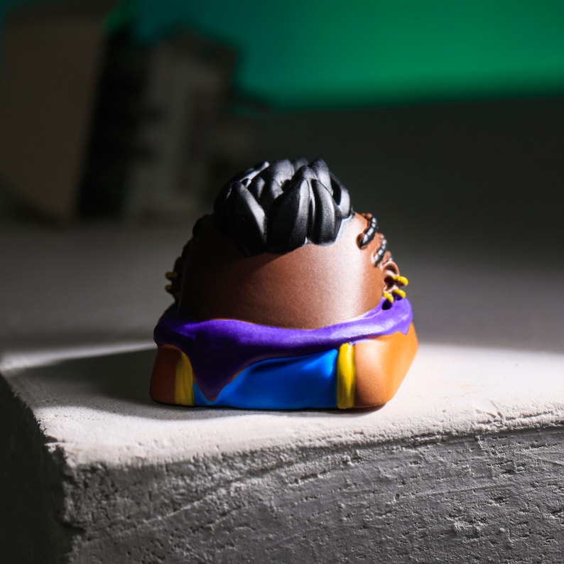 Pode incluir: Uma figura marrom e preta com um design roxo e azul. A figura tem uma superf&iacute;cie texturizada e uma pe&ccedil;a de cabelo preta.