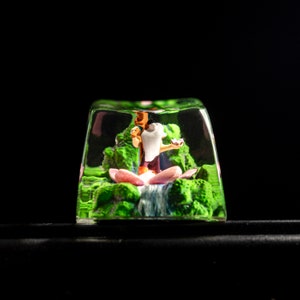May include: A clear resin keycap with a small figurine of a person sitting in a lotus flower. The figurine is surrounded by green foliage and a small waterfall.