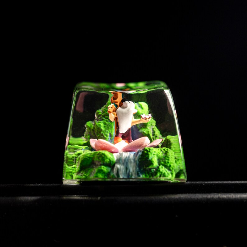 May include: A clear resin keycap with a small figurine of a person sitting in a lotus flower. The figurine is surrounded by green foliage and a small waterfall.