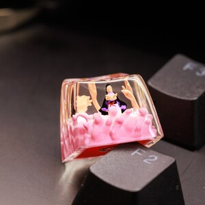Puede incluir: Un keycap de resina transparente con un dise&ntilde;o floral rosa y blanco. Una peque&ntilde;a figura de una persona con un atuendo morado y blanco est&aacute; dentro del keycap.