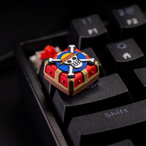 May include: A custom keycap featuring a red treasure chest design with gold accents and a blue circle with a skull and crossbones logo. The keycap is placed on a black keyboard, with the "Shift" key visible.