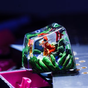 Puede incluir: Un keycap de resina transparente con una escena en miniatura en su interior. La escena presenta una peque&ntilde;a figura con armadura roja y dorada sosteniendo una espada, de pie en un paisaje verde y marr&oacute;n.