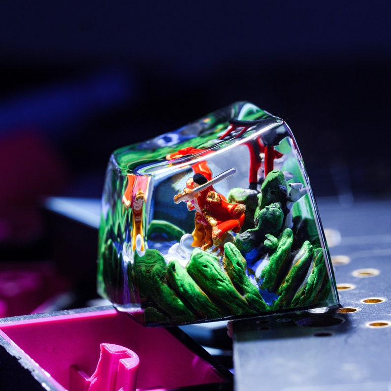 Puede incluir: Un keycap de resina transparente con una escena en miniatura en su interior. La escena presenta una peque&ntilde;a figura con armadura roja y dorada sosteniendo una espada, de pie en un paisaje verde y marr&oacute;n.