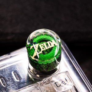 Puede incluir: Un tapón de botella redondo y transparente con la palabra "ZELDA" en letras doradas, sobre un diseño verde y negro. También se ve un pequeño gráfico de espada y escudo. El tapón está sobre un fondo oscuro.