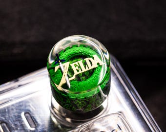May include: A clear, round bottle stopper featuring the word "ZELDA" in gold lettering, with a green and black design. A small sword and shield graphic is also visible. The stopper is set against a dark background.