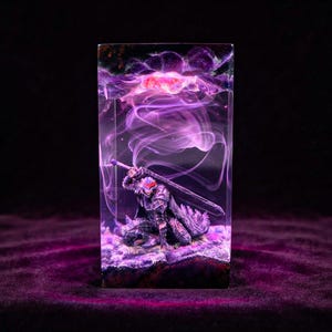 Pode incluir: Um bloco retangular de resina contendo uma figura em miniatura de um guerreiro com armadura escura segurando uma grande espada. O interior apresenta padrões de fumaça roxa e rosa e um efeito de luz brilhante no topo do bloco.