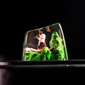 May include: A clear glass paperweight with a miniature scene inside. The scene features a small green forest with a brown and white figure standing in the middle.