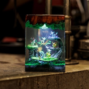 May include: A rectangular resin block lamp featuring a miniature forest diorama with multiple small creature figurines. The scene includes green mossy terrain, a brown tree structure, and internal LED lighting that illuminates the figures.