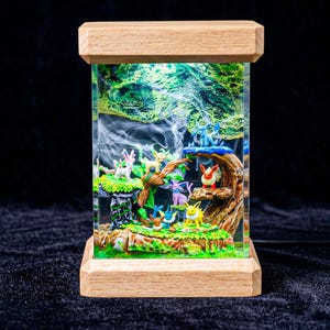 May include: A rectangular resin art piece with a miniature forest scene, illuminated from within. The base and top are made of light-coloured wood. The scene features trees, foliage, and several small, cartoon-like creatures in various colours.