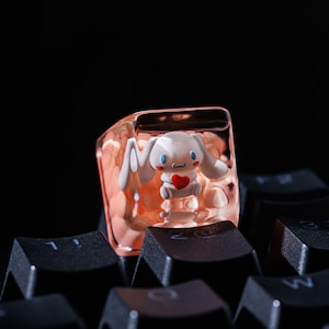 May include: A custom keycap with a white cartoon character holding a red heart, set within a translucent, peach-toned square. The keycap is placed on a black keyboard.