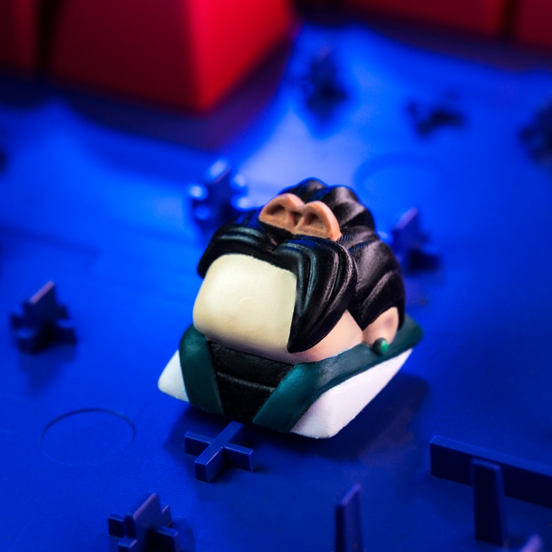 Pode incluir: Um keycap de resina verde, branco e preto com um personagem de desenho animado com bigode e uma brinco verde. O keycap est&aacute; sobre uma superf&iacute;cie azul.