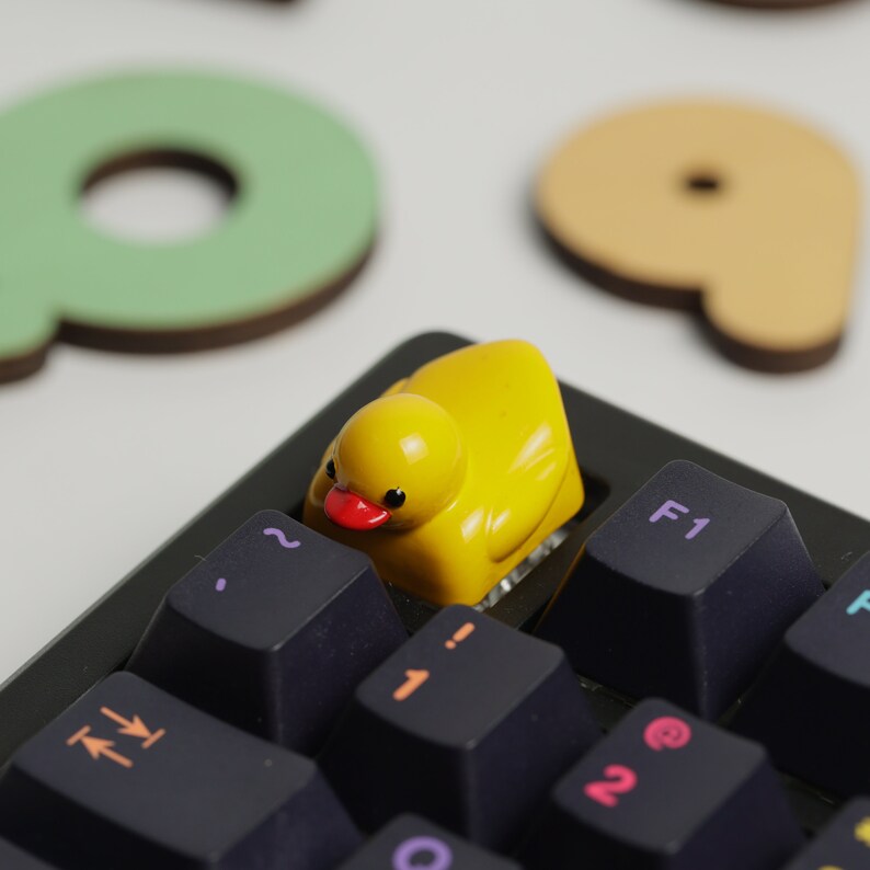 Puede incluir: Un tap&oacute;n de tecla de pato de goma amarillo se encuentra en un teclado mec&aacute;nico negro con etiquetas de teclas blancas. El teclado tiene teclas etiquetadas con letras, n&uacute;meros y s&iacute;mbolos. El tap&oacute;n de tecla de pato es una opci&oacute;n popular para jugadores y entusiastas de los teclados.