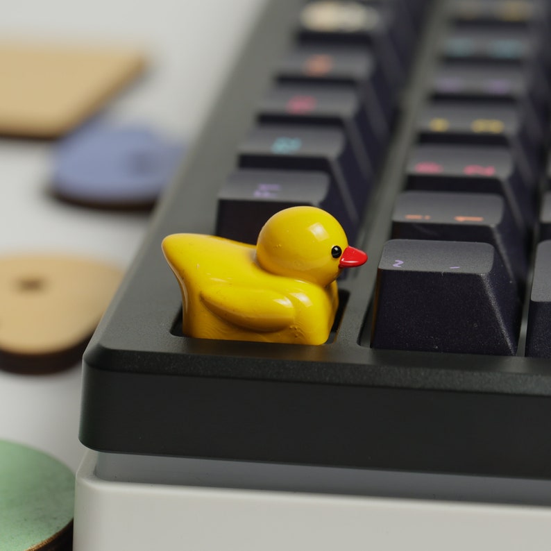 Puede incluir: Un tap&oacute;n de tecla de pato de goma amarillo con un pico rojo sobre un teclado negro.