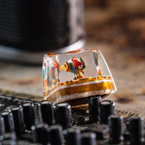 Puede incluir: Un tap&oacute;n de tecla de resina transparente con una peque&ntilde;a figura de un personaje de dibujos animados de colores en su interior. La figura lleva un sombrero rojo y un atuendo amarillo y azul. El tap&oacute;n de tecla est&aacute; sobre una superficie negra.