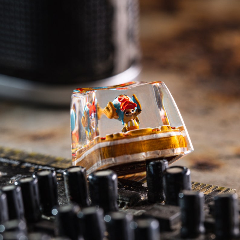 Puede incluir: Un tap&oacute;n de tecla de resina transparente con una peque&ntilde;a figura de un personaje de dibujos animados de colores en su interior. La figura lleva un sombrero rojo y un atuendo amarillo y azul. El tap&oacute;n de tecla est&aacute; sobre una superficie negra.