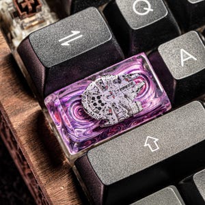 Puede incluir: Un tap&oacute;n de tecla de resina morado y rosa con un dise&ntilde;o de nave espacial Millennium Falcon blanco. El tap&oacute;n de tecla est&aacute; en un teclado negro.